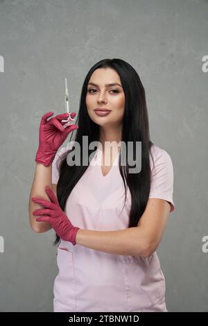 Estetista femminile sicura di sé che indossa uniforme rosa in piedi con la siringa all'interno. Ritratto di una bella donna caucasica che indossa guanti che tiene la siringa e sorride in clinica. Concetto di cura della bellezza. Foto Stock