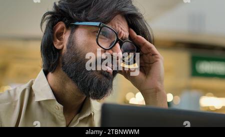 Indiano barbuto stressato uomo in occhiali che lavora in ufficio con portatile uomo d'affari arabo triste con problemi di business mal di testa soffre di Foto Stock