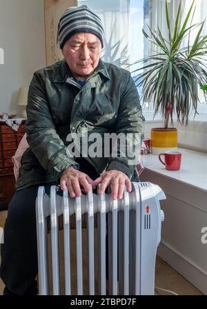 Un uomo anziano completamente vestito nel soggiorno, seduto accanto a un riscaldatore elettrico portatile, che cerca di tenersi al caldo durante il freddo inverno. Foto Stock