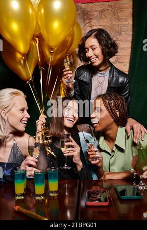 fidanzate multietniche alla moda con champagne sorridente vicino a cocktail e palloncini dorati, compleanno Foto Stock