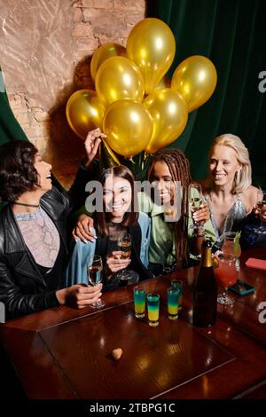 felice ed elegante donna multietnica con champagne vicino a palloncini dorati e cocktail al bar, compleanno Foto Stock