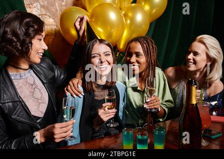 gioiose amiche multietniche con champagne che festeggia il compleanno vicino a palloncini dorati nel bar Foto Stock