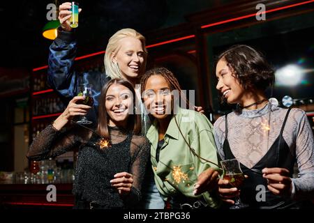 amiche multietniche felici e alla moda che brindano con cocktail e tengono scintillanti scintillanti al bar Foto Stock