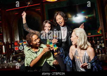 eccitate amiche multietniche con scintillanti che si bevono bicchieri da cocktail al bar, feste vivaci Foto Stock