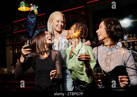 amiche multietniche felici ed eleganti con cocktail e scintillanti festivi durante la festa al bar Foto Stock