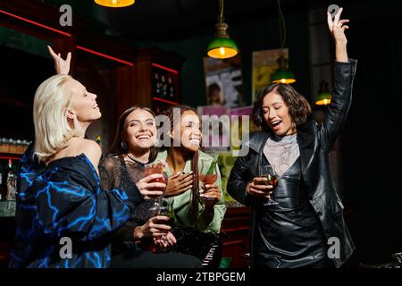 fidanzate multietniche affascinanti e felici che ballano con bicchieri da cocktail al bar, feste glamour Foto Stock