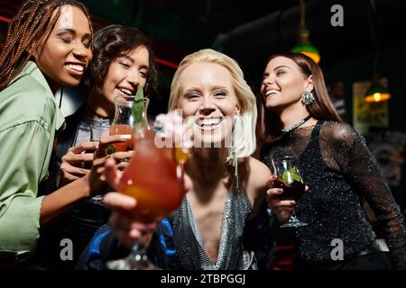 felice donna bionda con un bicchiere di cocktail sorridente alla macchina fotografica vicino a fidanzate multietniche al bar Foto Stock