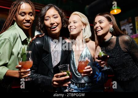 giovani ed eleganti fidanzate multietniche con bicchieri da cocktail sorridenti al bar, feste di addio al nubilato Foto Stock