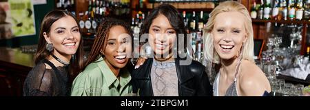 amiche multietniche gioiose ed eleganti con bicchieri da cocktail che guardano la macchina fotografica al bar e allo striscione Foto Stock