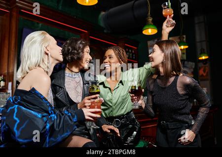 eccitata donna afroamericana che brinda con un cocktail vicino a eleganti amiche multietniche al bar Foto Stock