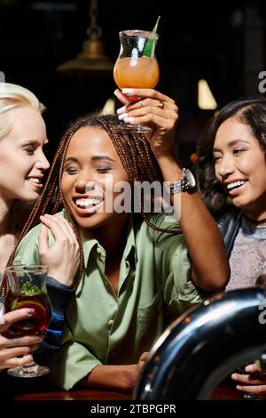 allegra donna afroamericana che brinda con un bicchiere da cocktail vicino a eleganti amiche multietniche Foto Stock