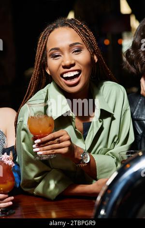 donna afro-americana eccitata e alla moda con un bicchiere da cocktail che ride vicino alle amiche nel bar Foto Stock