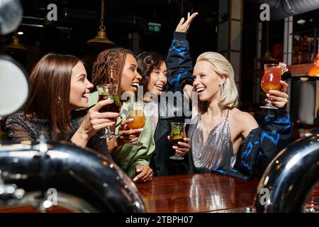 donna eccitata che tosta con cocktail e ha alzato la mano durante una festa al bar con amiche multietniche Foto Stock