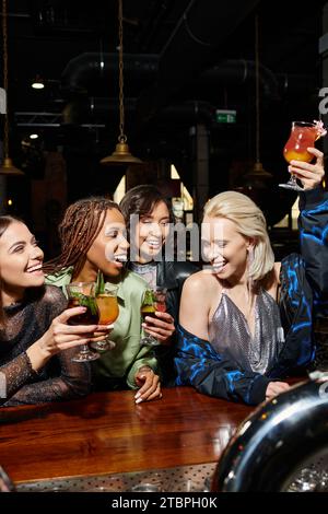 eccitata donna bionda che brinda con un cocktail durante la festa di nubilato con amiche multietniche al bar Foto Stock