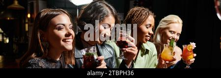 amiche multiculturali felici ed eleganti che tengono deliziosi cocktail nel bar notturno, banner Foto Stock