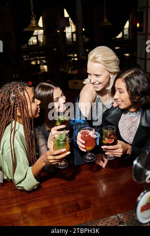 fidanzate multietniche alla moda con bicchieri da cocktail che parlano al bar, atmosfera elegante Foto Stock