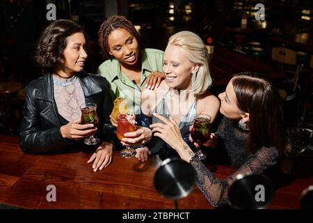 amiche multietniche sorridenti ed eleganti con cocktail che parlano al bar, tempo libero notturno Foto Stock