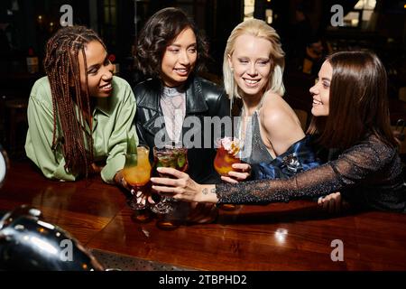 amiche multietniche gioiose ed eleganti che bevono bicchieri da cocktail al bar, vita notturna moderna Foto Stock