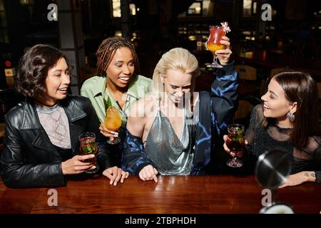 eccitata donna bionda che tosta con un bicchiere da cocktail vicino a eleganti amiche multietniche al bar Foto Stock