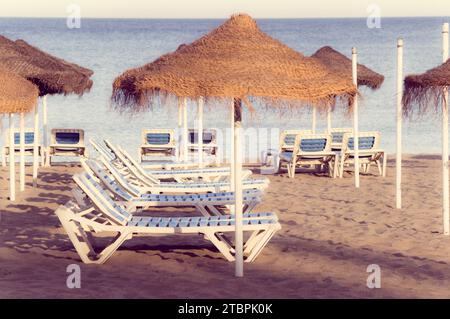 Un idilliaco scenario balneare con due sedie a sdraio e due grandi ombrelloni in paglia disposti nella sabbia Foto Stock