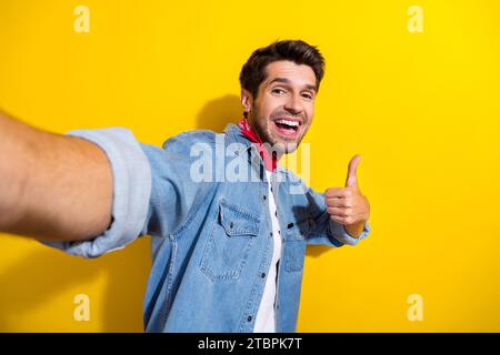 Ritratto fotografico di un giovane attraente scatta selfie mostra il pollice in alto vestiti eleganti in denim isolati su sfondo di colore giallo Foto Stock