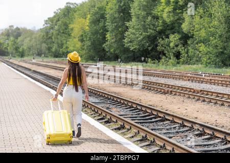 Un'appassionata turista, vestito con eleganti abiti gialli, aspetta il suo tanto atteso viaggio, tenendo in mano una valigia. Una donna cammina con una valigia sul Foto Stock