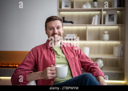 Felice uomo sorridente seduto in un accogliente appartamento con una tazza di tè, bevendo, riposando, guardando la macchina fotografica Foto Stock