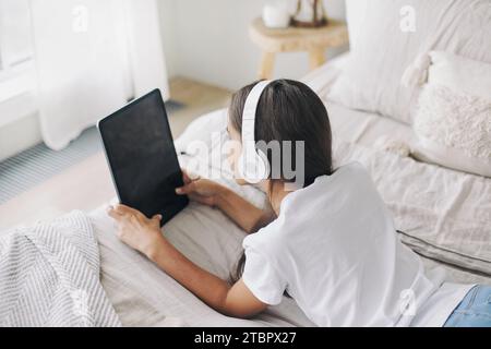 Adorabile ragazza pre-adolescente dei 12 anni con cuffie wireless sdraiate sul letto con un tablet digitale. Giovani generazioni e abuso di tecnologia moderna, uso di gadget per Foto Stock