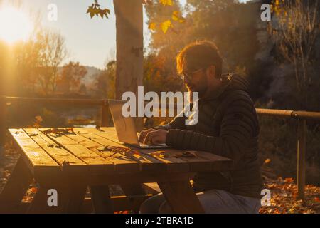 Un uomo da solo che lavora su un computer portatile all'aperto seduto su un tavolo di legno al parco durante un autunno dorato con un incredibile tramonto. Persone e piccole imprese o tecnologia nomade digitale che si spostano online Foto Stock