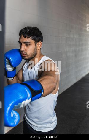 Verticale foto uomo giovane adulto corsa mista pugilato con borsa e guanti blu, in palestra. Copia spazio. Sport, attività ricreative. Foto Stock