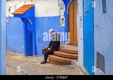 Uomo musulmano che indossa la tradizionale djellaba nel vicolo con case e porte blu nella medina / centro storico della città di Chefchaouen / Chaouen, Marocco Foto Stock