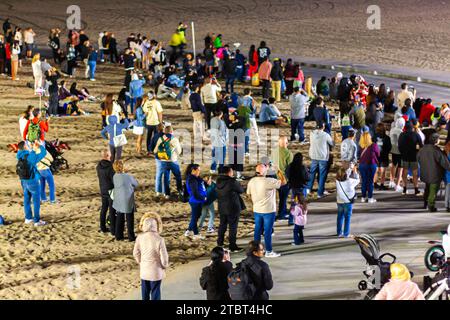 Una folla di persone in piedi sulla spiaggia di notte guardando il cielo guardando uno spettacolo di droni Foto Stock