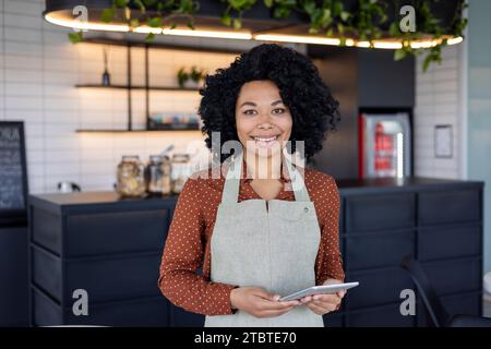 Ritratto cameriera di una piccola azienda proprietaria di un caffè, donna in grembiule sorride e guarda la fotocamera con un tablet in mano, un dipendente di un bar di un ristorante vicino a un bancone del caffè. Foto Stock