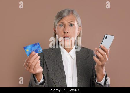 Donna d'affari scioccata con carta di credito e smartphone su sfondo marrone. Attenzione - frode Foto Stock