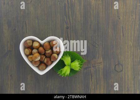 Nocciole fresche non sgusciate in un piatto a forma di cuore con foglie verdi in un piano in alto stendere la vita su un tavolo di legno con copyspace Foto Stock