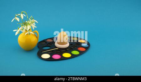Decorazione con uovo di Pasqua con set di vernici acquerello in paletta, accanto a una caraffa gialla con freschi fiori bianchi primaverili. Studio STILL Life On Foto Stock