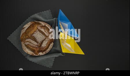 Pane di pane posto su un tavolo nero vicino bandiera Ucraina colori barche origami, concetto di esportazioni di grano e crisi alimentare globale, vista dall'alto con spazio copia Foto Stock