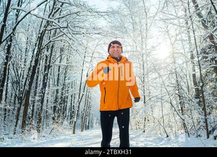 Corridore sorridente di mezza età vestito da giacca antivento arancione brillante, che corre lungo la pittoresca foresta innevata. Persone sportive attive e vincenti Foto Stock