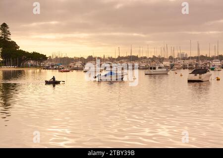 Uomo che canoa in barca sotto la luce dorata davanti a barche e yacht al Freshwater Bay Yacht Club sul fiume Swan, Perth, Australia Occidentale. Foto Stock