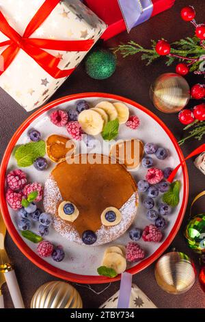 Divertenti e carine frittelle di volpe natalizie su un piatto rosso bianco decorato con bacche fresche, frutta e zucchero in polvere, colazione di Natale per bambini Foto Stock