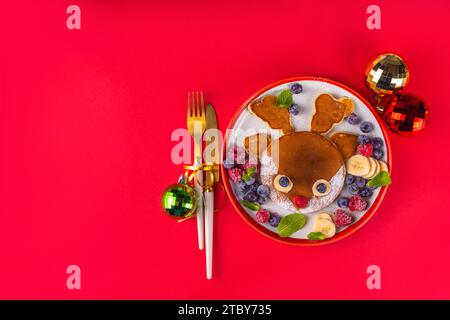 Divertenti e carine frittelle di renna natalizie su un piatto rosso bianco decorato con bacche fresche, frutta e zucchero in polvere, colazione di Natale per bambini Foto Stock