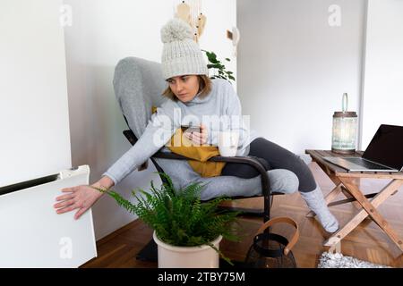 Una donna con abiti caldi e smartphone siede vicino a un radiatore di riscaldamento. Bassa temperatura in casa. Concetto di crisi energetica.. Foto Stock