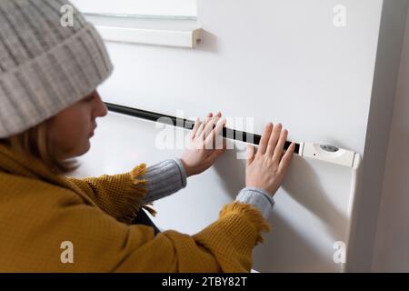 Una donna in abiti caldi tiene la mano sul riscaldatore. Bassa temperatura di riscaldamento in casa. Concetto di crisi energetica.. Foto Stock