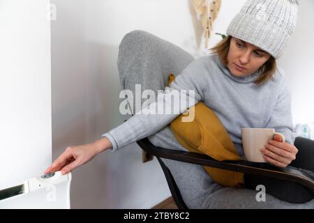 Una donna con abiti caldi tiene la mano sul termostato del riscaldatore. Bassa temperatura di riscaldamento in casa. Concetto di crisi energetica.. Foto Stock