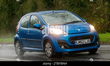 Milton Keynes,Bucks,Regno Unito - 9 dicembre 2023: 2012 Peugeot blu 107 guida su una strada bagnata, sotto la pioggia con i fari accesi Foto Stock