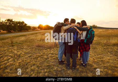 Squadra di amici felici con zaini che si avvolgono e si abbracciano nel campo durante il tour escursionistico Foto Stock