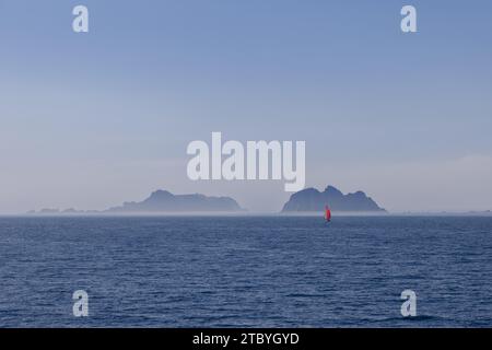 Una barca a vela solitaria con una vivace vela rossa, naviga con grazia nel tranquillo Mare del Nord vicino alle isole Lofoten, con maestose creste montuose velate di nebbia Foto Stock