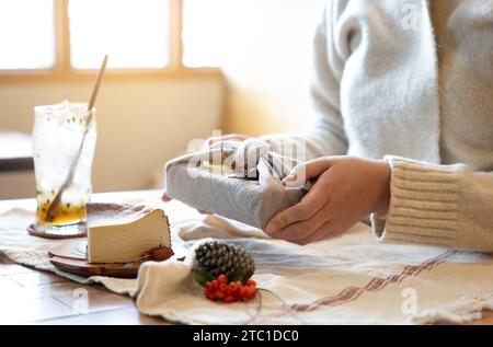 Mano di donna che avvolge un regalo con un vecchio panno vintage alla finestra con la luce del sole. Foto sfocata con coni di pino e materiali naturali per decorati Foto Stock