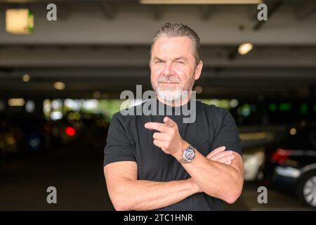 L'attraente uomo dai capelli grigi con una maglietta nera indossa un orologio elegante e punta verso la fotocamera, di fronte a un parcheggio Foto Stock
