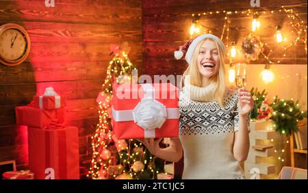 Donna bionda felice nel cappello rosso di Babbo Natale che tiene un regalo regalo in una fande e una vedova champagne in un altro al bell'interno decorato Foto Stock